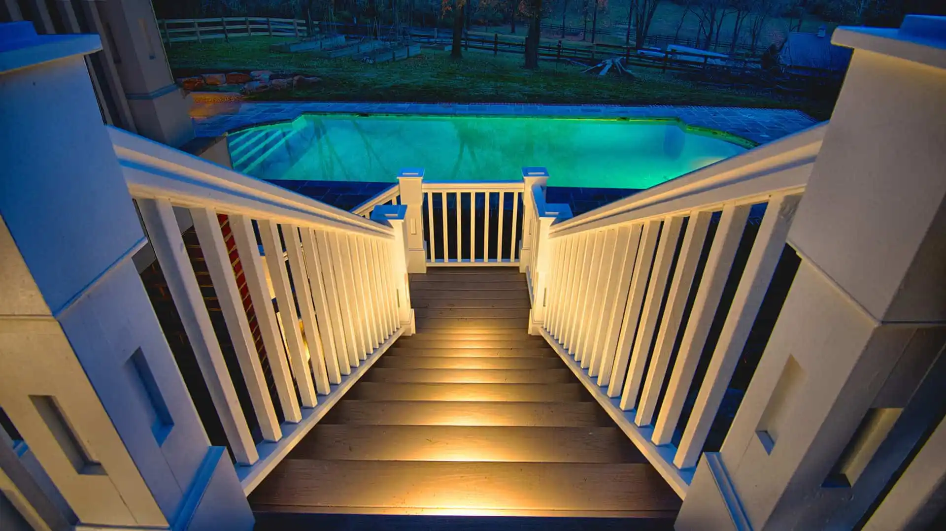 Wide-angle view of illuminated wooden stairs leading down to a glowing outdoor pool at night. Expert outdoor living design in Delaware County, PA enhances the serene ambiance, with trees and a fence in the background creating an inviting atmosphere.