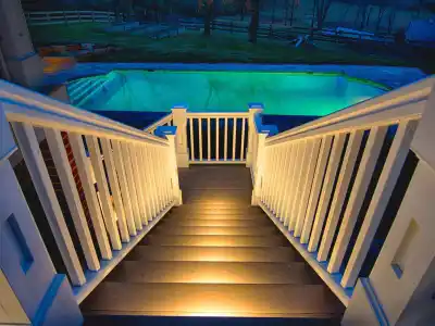 Wide-angle view of illuminated wooden stairs leading down to a glowing outdoor pool at night. Expert outdoor living design in Delaware County, PA enhances the serene ambiance, with trees and a fence in the background creating an inviting atmosphere.