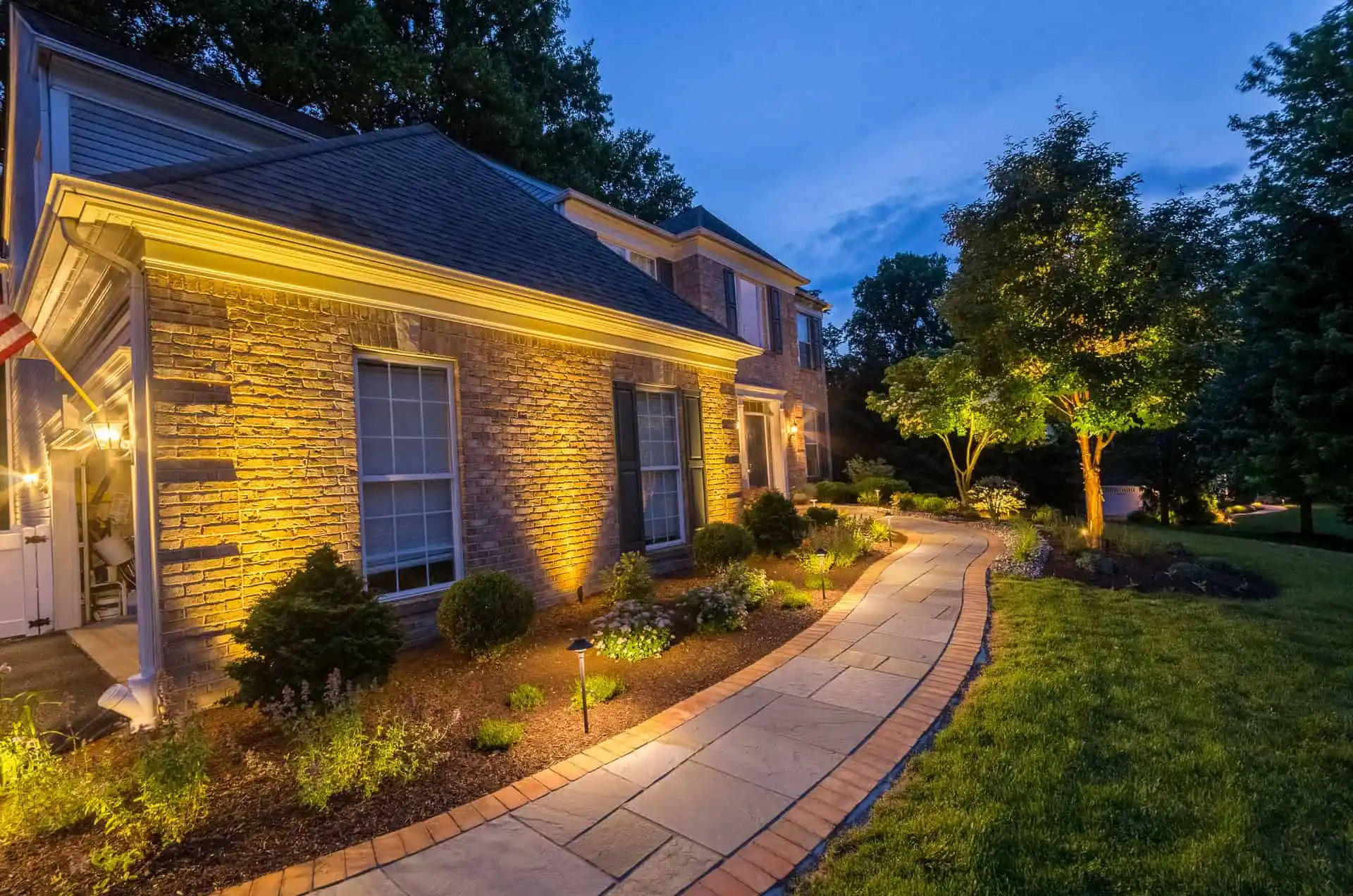 A stone house with large windows is illuminated by warm outdoor lights at dusk. A curved walkway bordered by plants and bushes leads through a well-kept yard with trees and grass.