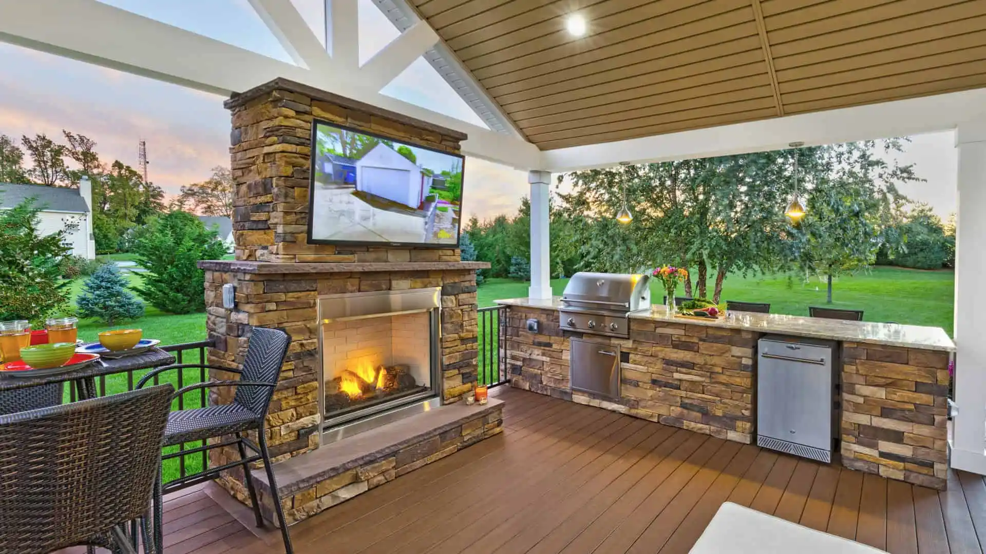 Experience the charm of outdoor living in Delaware County, PA, with a stunning patio featuring a stone fireplace and mounted TV. Enjoy grilling on the kitchen countertop equipped with a sink and mini fridge. Relax on wicker chairs as you take in the sunset over lush trees and lawn.
