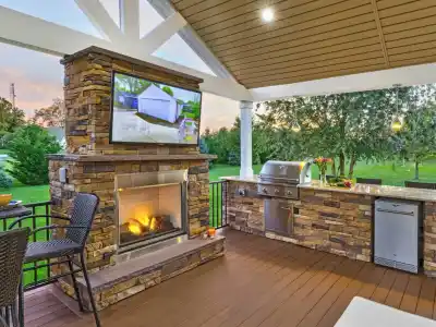 Experience the charm of outdoor living in Delaware County, PA, with a stunning patio featuring a stone fireplace and mounted TV. Enjoy grilling on the kitchen countertop equipped with a sink and mini fridge. Relax on wicker chairs as you take in the sunset over lush trees and lawn.