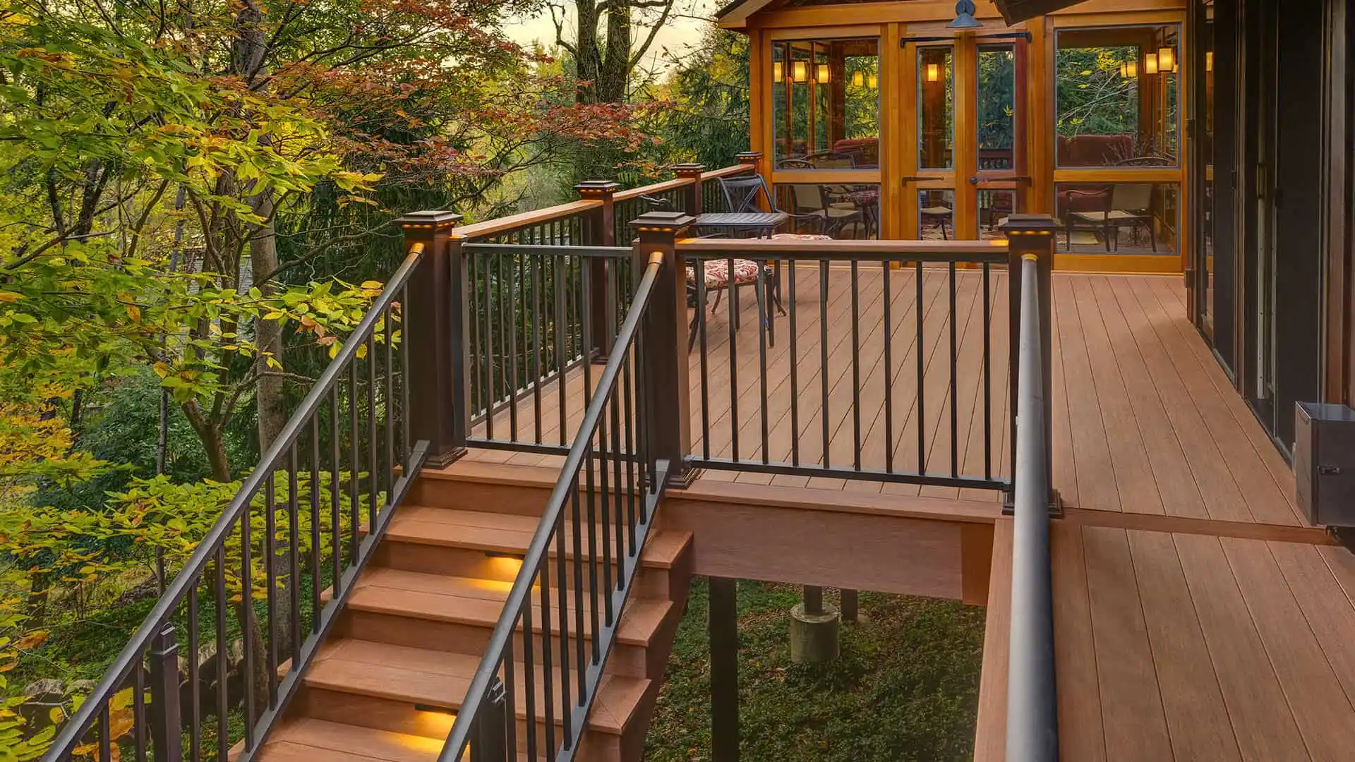 A wooden deck with illuminated stairs and metal railings overlooks a lush green forest in Delaware County, PA. Attached to a house with large windows, it offers an inviting space for outdoor living and seating to enjoy nature. Explore local hardscaping services to enhance your retreat.