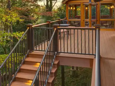 A wooden deck with illuminated stairs and metal railings overlooks a lush green forest in Delaware County, PA. Attached to a house with large windows, it offers an inviting space for outdoor living and seating to enjoy nature. Explore local hardscaping services to enhance your retreat.