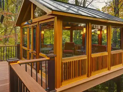 A cozy wooden sunroom with large windows extends from a house, surrounded by lush green trees. The interior features comfortable seating and warm lighting, making it a perfect outdoor living retreat in Delaware County, PA.
