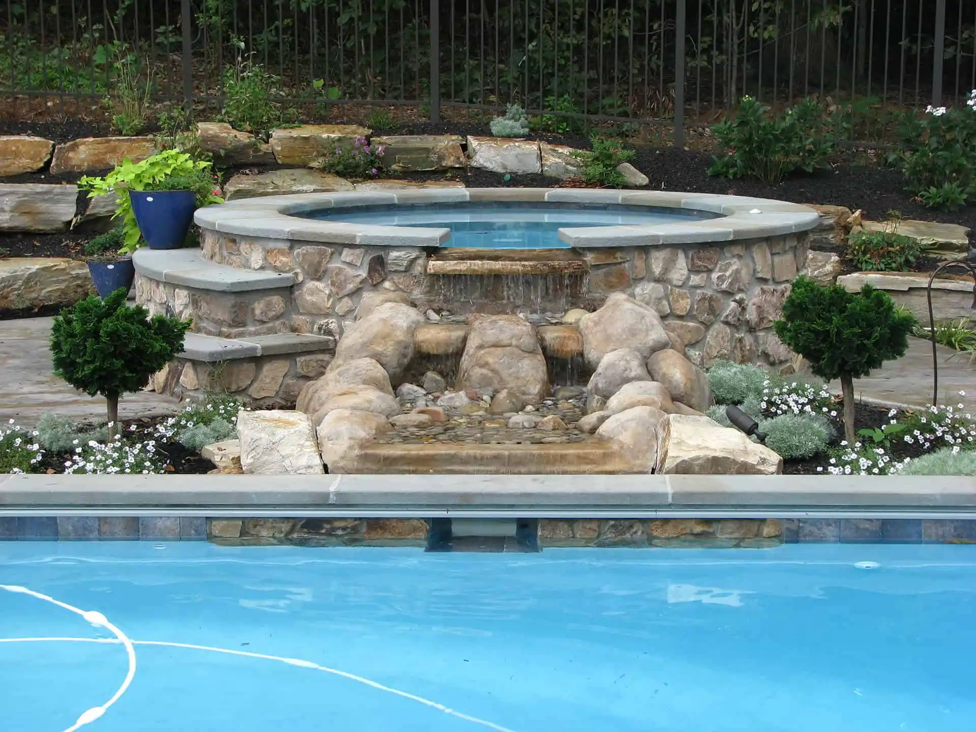A stone hot tub with a waterfall feature cascades into a pool below, surrounded by plants, small trees, and decorative rocks in a landscaped backyard.
