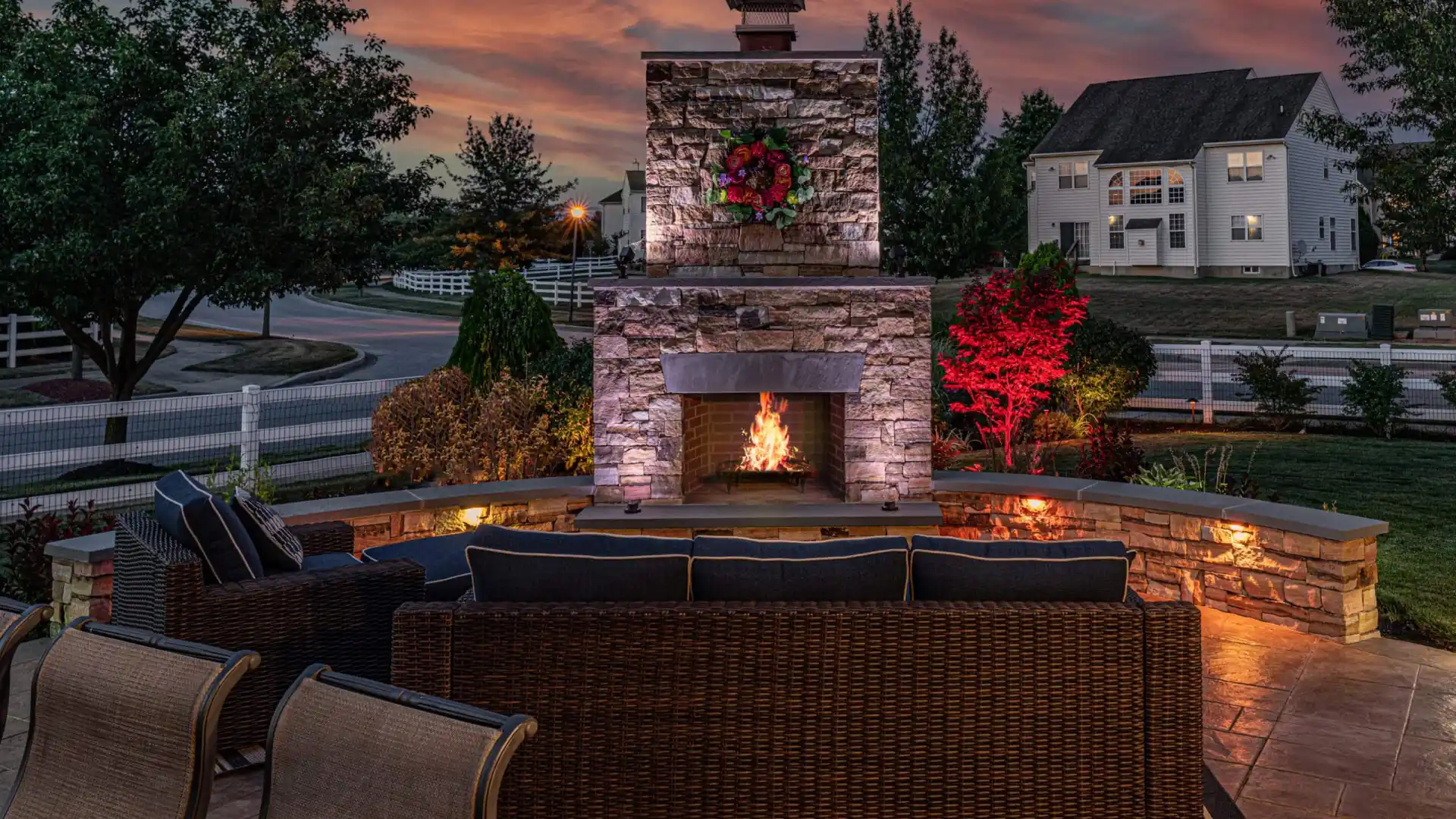 An inviting outdoor patio in Delaware County, PA showcases expert hardscaping services with a lit stone fireplace, wicker seating, and soft lighting against a vibrant sunset. Red and green plants surround the area, while a large house with glowing windows completes this outdoor living oasis.