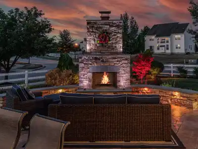 An inviting outdoor patio in Delaware County, PA showcases expert hardscaping services with a lit stone fireplace, wicker seating, and soft lighting against a vibrant sunset. Red and green plants surround the area, while a large house with glowing windows completes this outdoor living oasis.