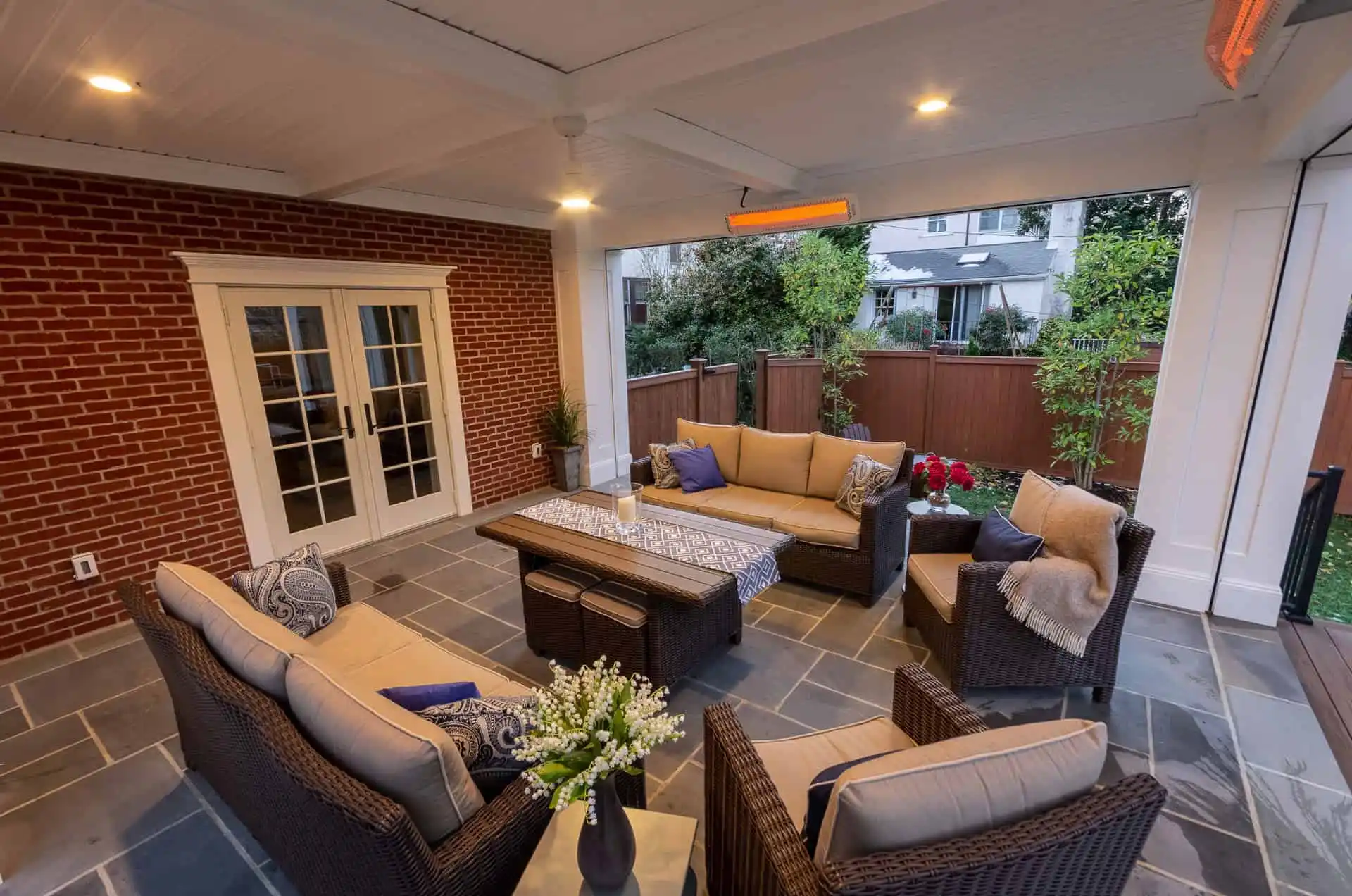 A covered patio with wicker furniture, beige cushions, a central coffee table, plants, decorative pillows, and soft lighting. French doors lead inside; a wooden fence and houses are visible outside.