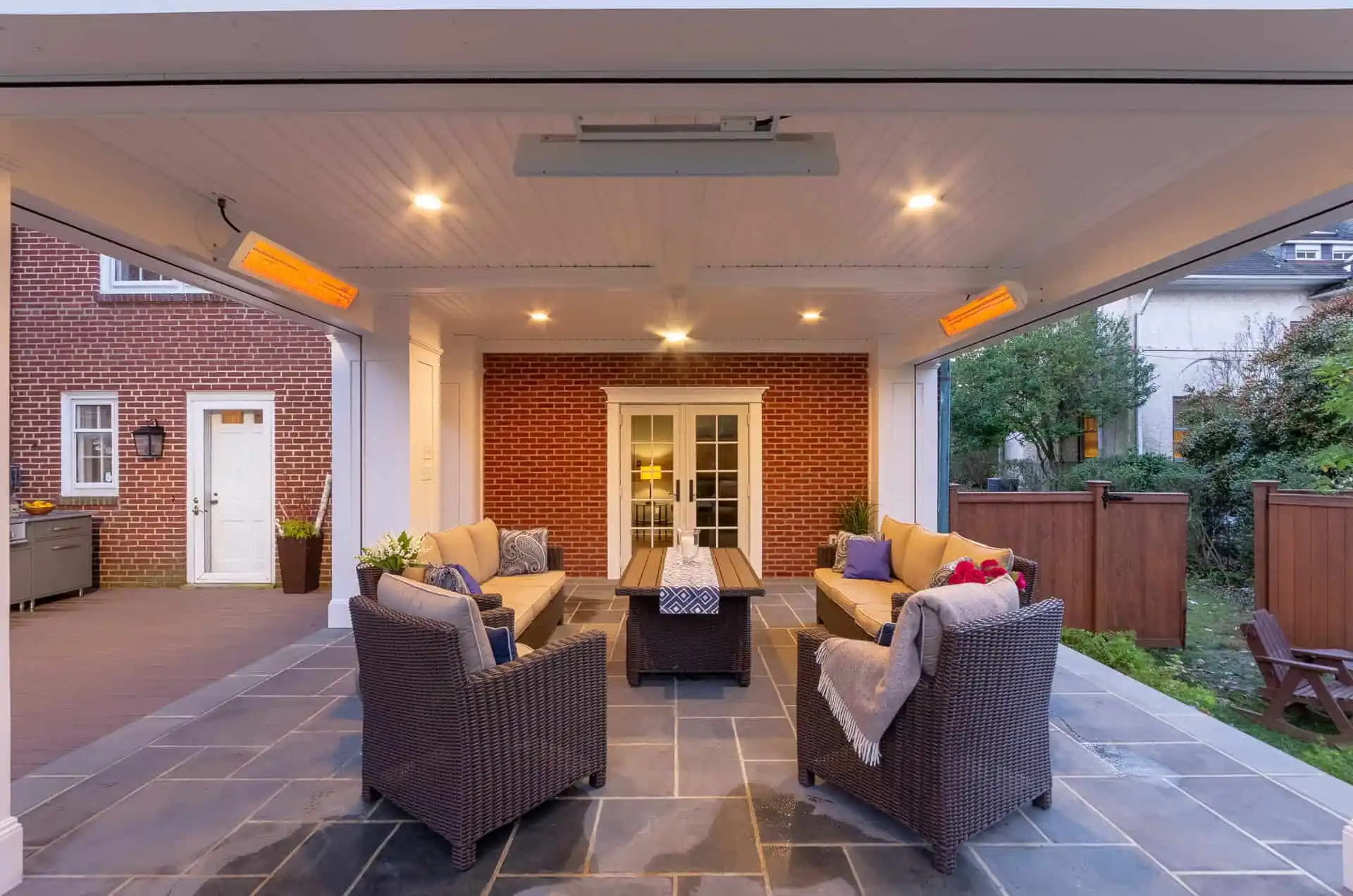 A covered outdoor patio with wicker furniture, cushioned chairs, a coffee table, ceiling heaters, and recessed lighting, set on gray stone tiles against a brick house with French doors.