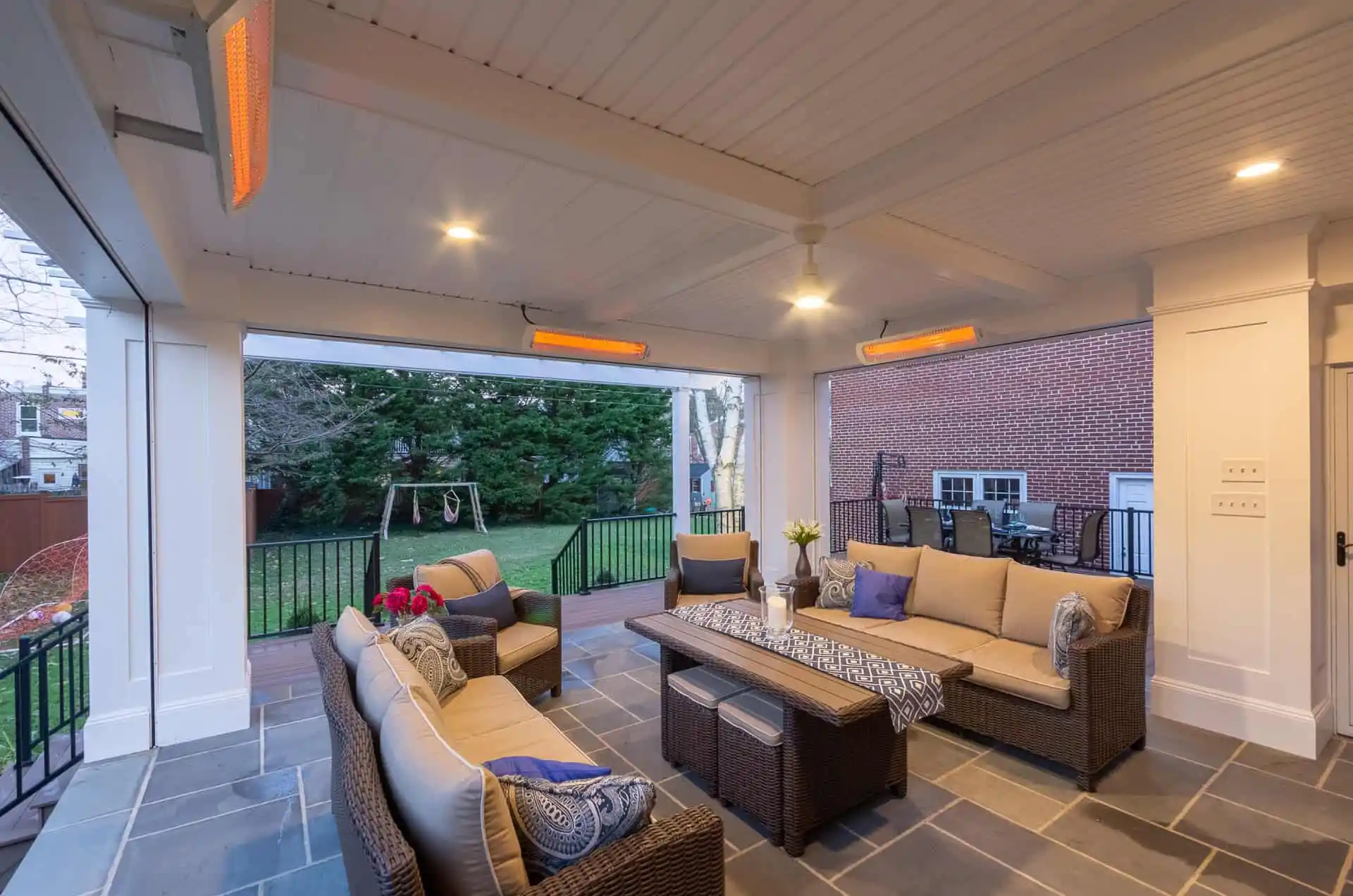A covered patio with wicker furniture, beige cushions, a coffee table, ceiling fans, and wall heaters, overlooking a fenced backyard with a swing set and trees. Outdoor dining area is visible in the background.