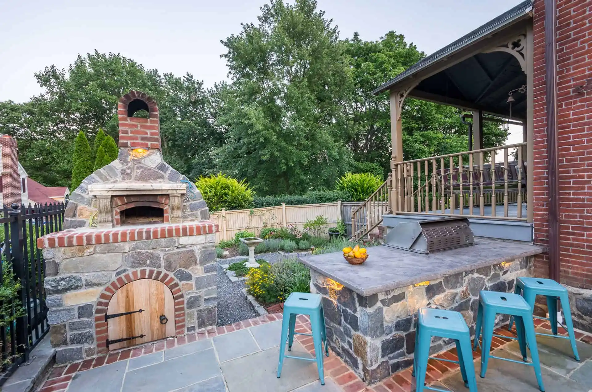 Stone outdoor kitchen with a wood-fired pizza oven, a countertop with a grill, four blue stools, and a bowl of lemons. A porch and lush green trees are visible in the background.