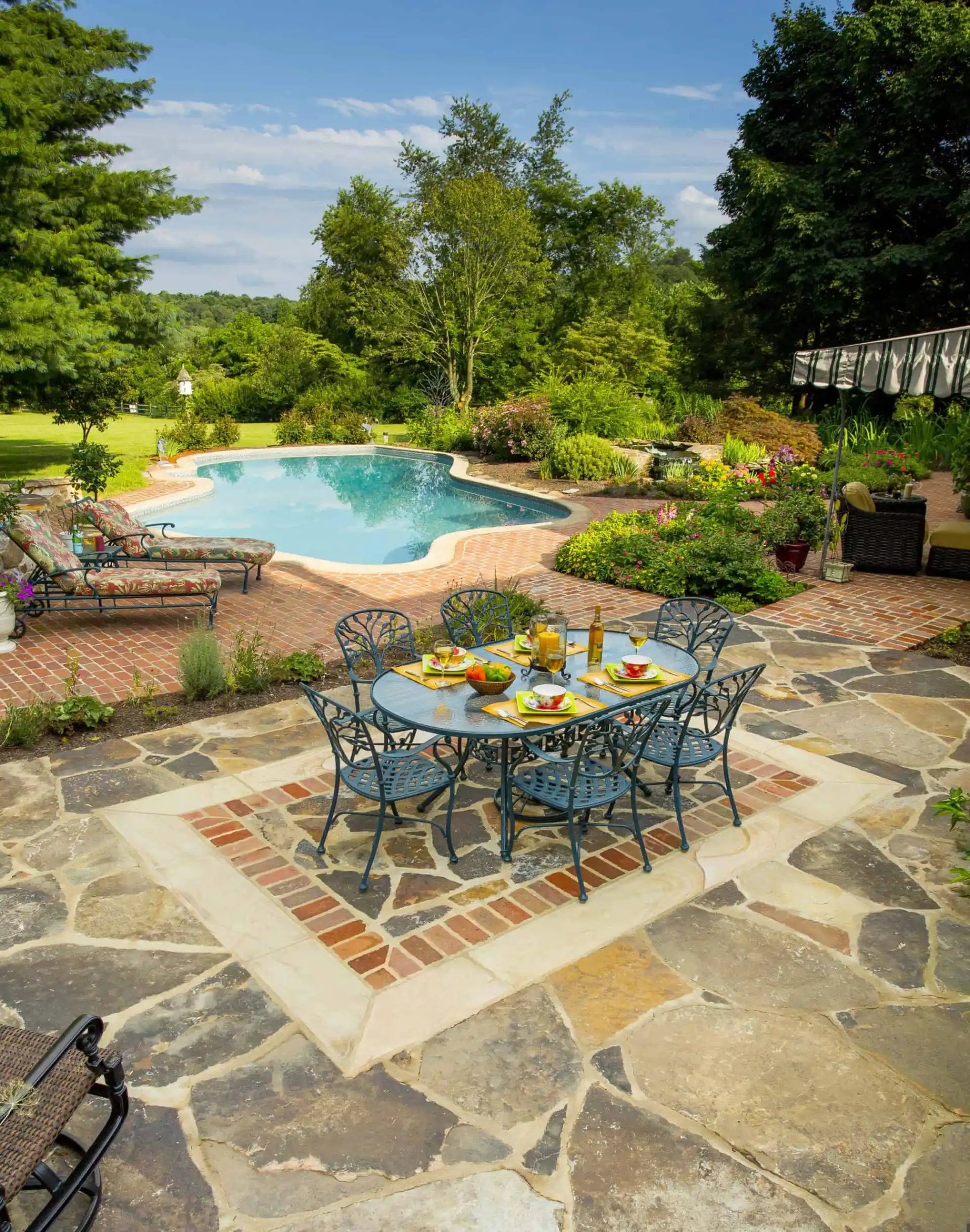 A backyard patio with a metal dining table set for four, surrounded by stone and brick paving, overlooking a landscaped garden and a kidney-shaped swimming pool. Lush greenery and trees fill the background.