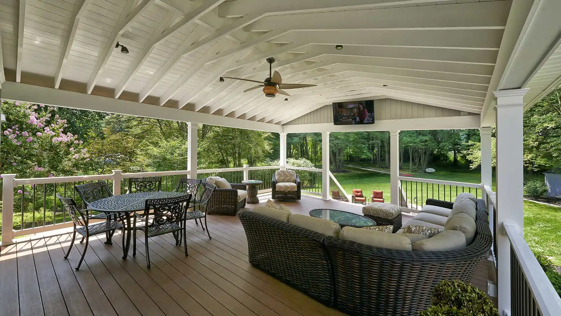 This outdoor living space in Delaware County, PA, boasts a covered patio with wooden flooring and a vaulted ceiling. It features a ceiling fan, TV, wicker sofas, cushioned armchairs, and a round metal table with chairs—all set against a natural backdrop of trees and grass.