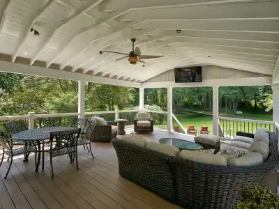 This outdoor living space in Delaware County, PA, boasts a covered patio with wooden flooring and a vaulted ceiling. It features a ceiling fan, TV, wicker sofas, cushioned armchairs, and a round metal table with chairs—all set against a natural backdrop of trees and grass.