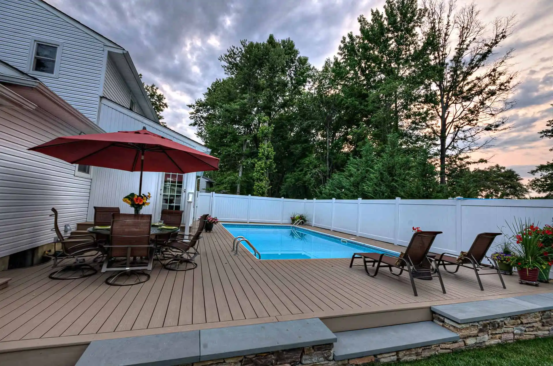 A backyard with a rectangular swimming pool, wooden deck, lounge chairs, a dining table with chairs, and a large red umbrella, surrounded by a white privacy fence and tall green trees.