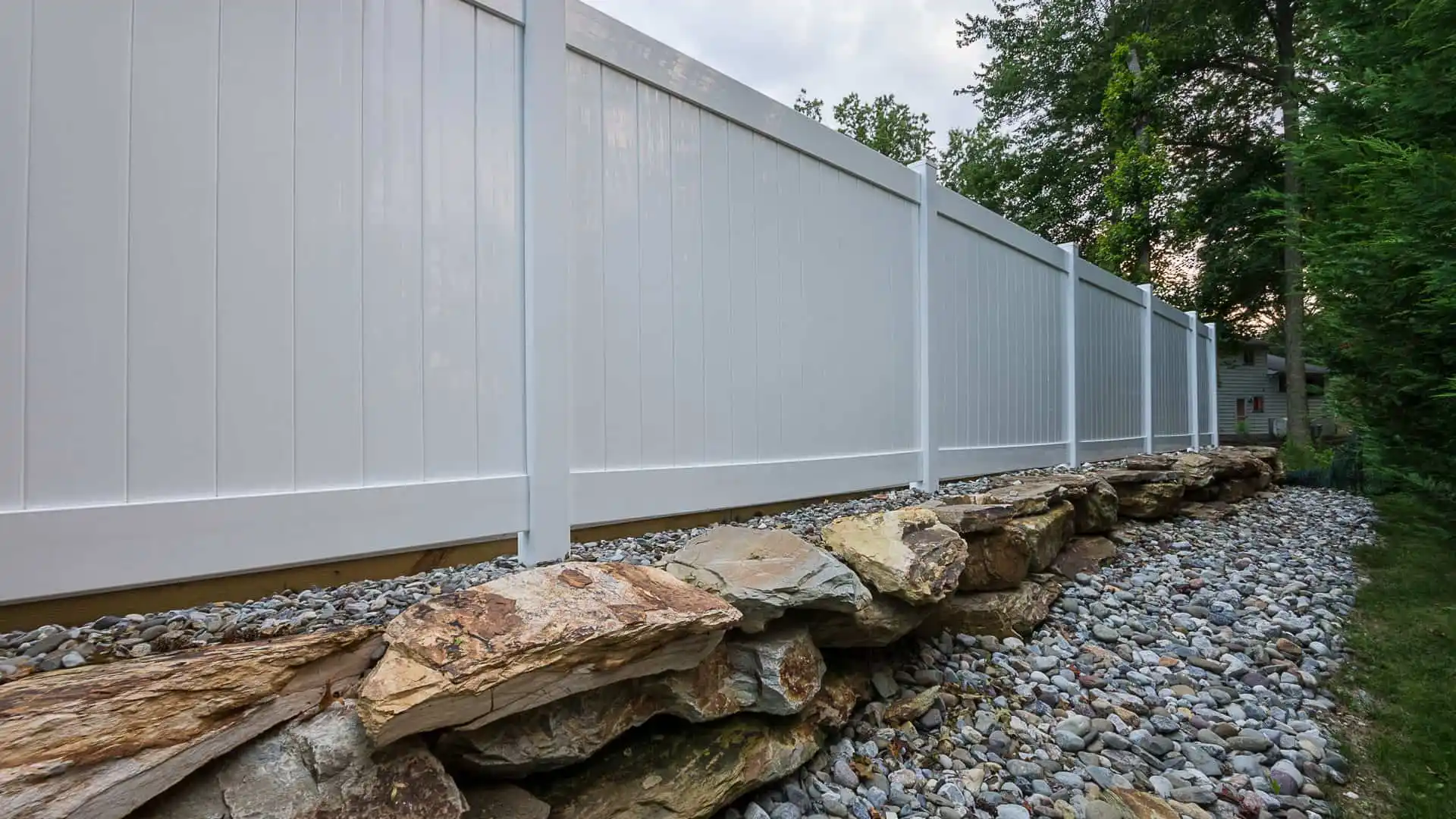 A long white vinyl fence atop a rocky retaining wall, surrounded by small gray stones, defines this outdoor living space in Delaware County, PA. Trees and foliage peek through under a cloudy sky, showcasing expert landscaping services.