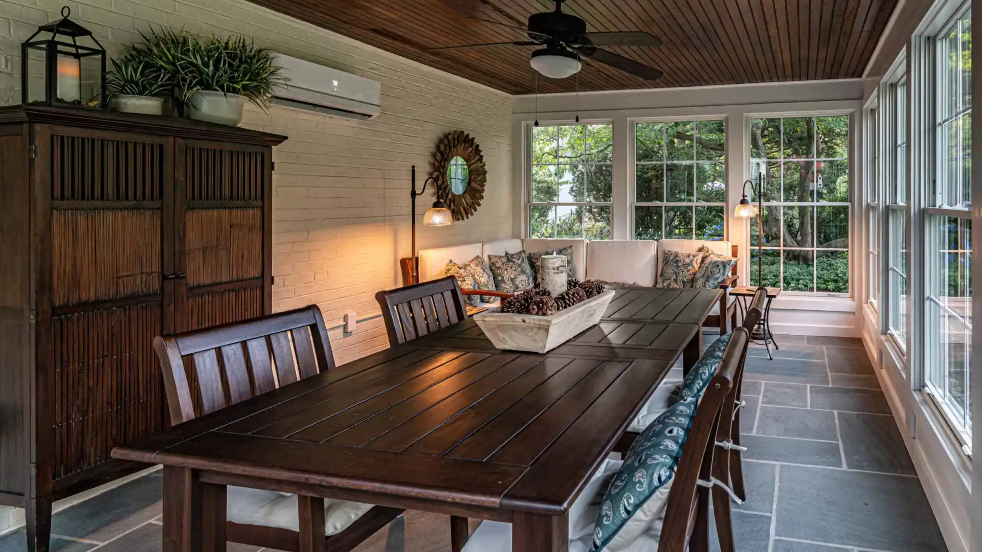 Sunroom with a dining table and six chairs, a cushioned seating area, wooden ceiling, and large windows showcasing the lush greenery created by outdoor living Delaware County PA. Decor includes plants, a mirror, a ceiling fan, and a wicker tray on the table.