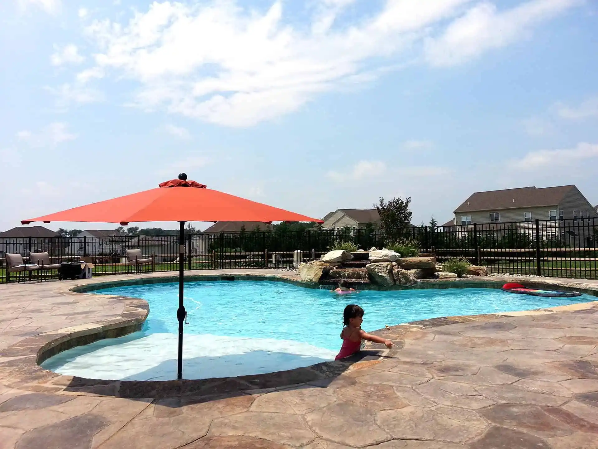 A young child stands in a backyard pool near a rock waterfall. A large red umbrella shades a shallow section of the pool. Patio chairs and houses are visible in the background under a partly cloudy sky.