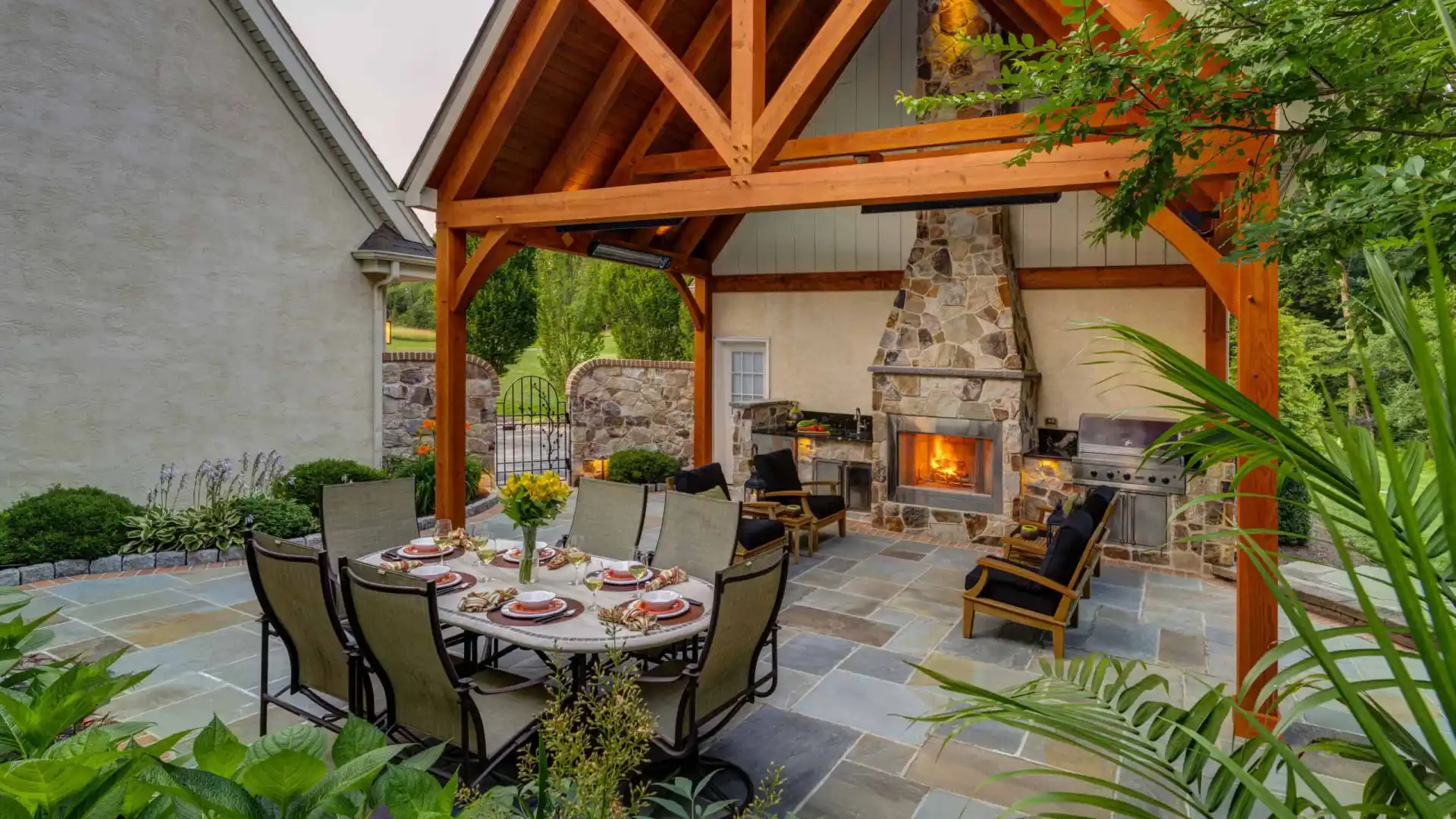 Outdoor living in Delaware County, PA, shines with a stone fireplace under a wooden pergola. A dining table for six is elegantly set, complemented by a nearby barbecue grill surrounded by lush greenery and trees. Rustic stone flooring showcases expert hardscaping services.