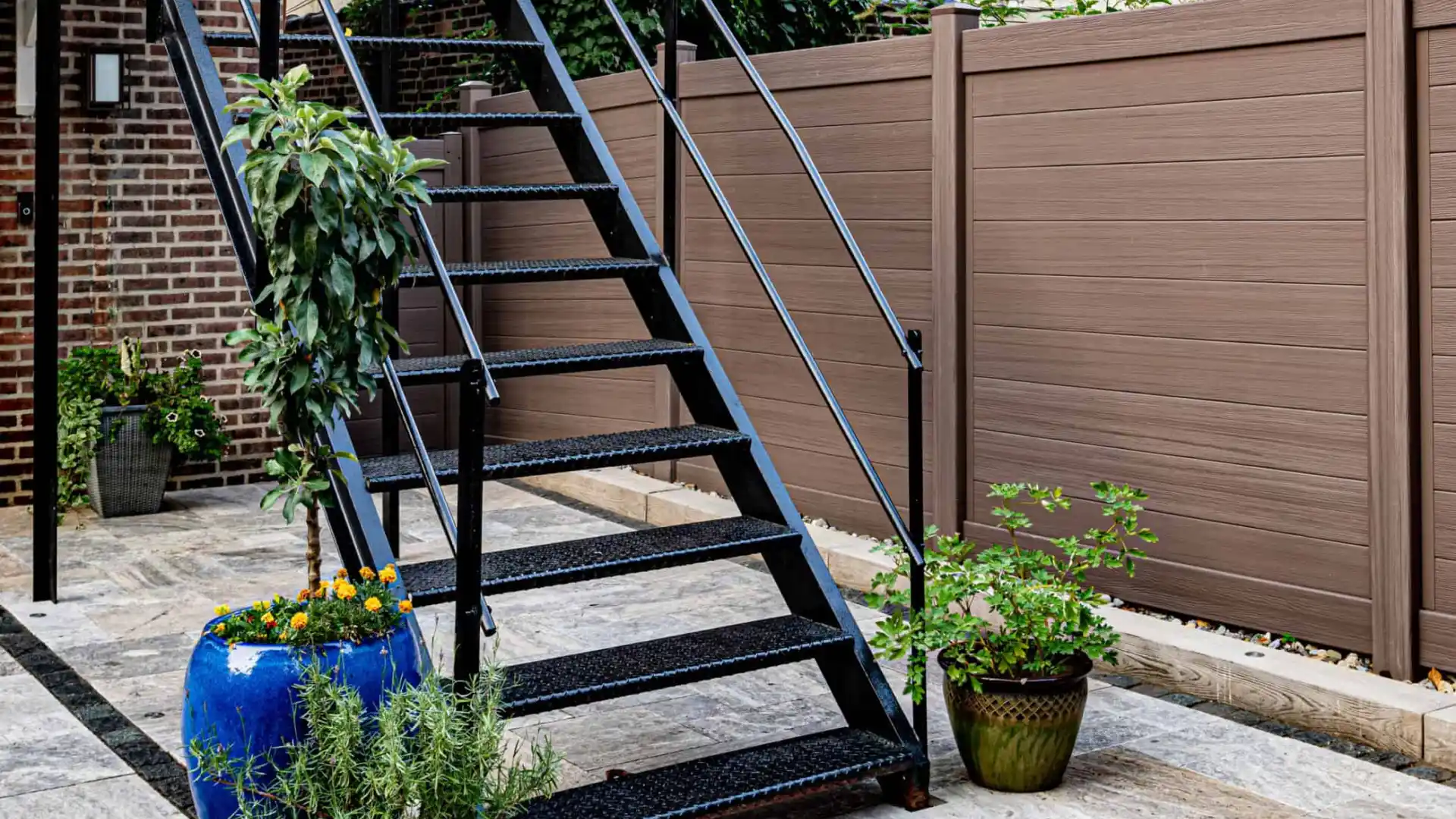 The outdoor scene features metal stairs ascending to a building, with potted plants artfully arranged at the base, including a tall plant in a large blue pot. A brown fence lines the tiled ground as nearby buildings stand visible, showcasing superb hardscaping services in Delaware County PA.