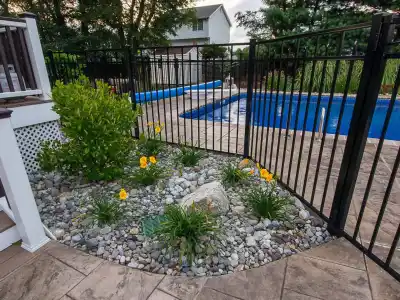 A fenced-in backyard pool area with a paved path showcases the epitome of outdoor living in Delaware County, PA. Yellow flowers and green shrubs thrive in a bed of small rocks. Stairs with a white railing lead up to a deck, framed by trees and a house in the background.