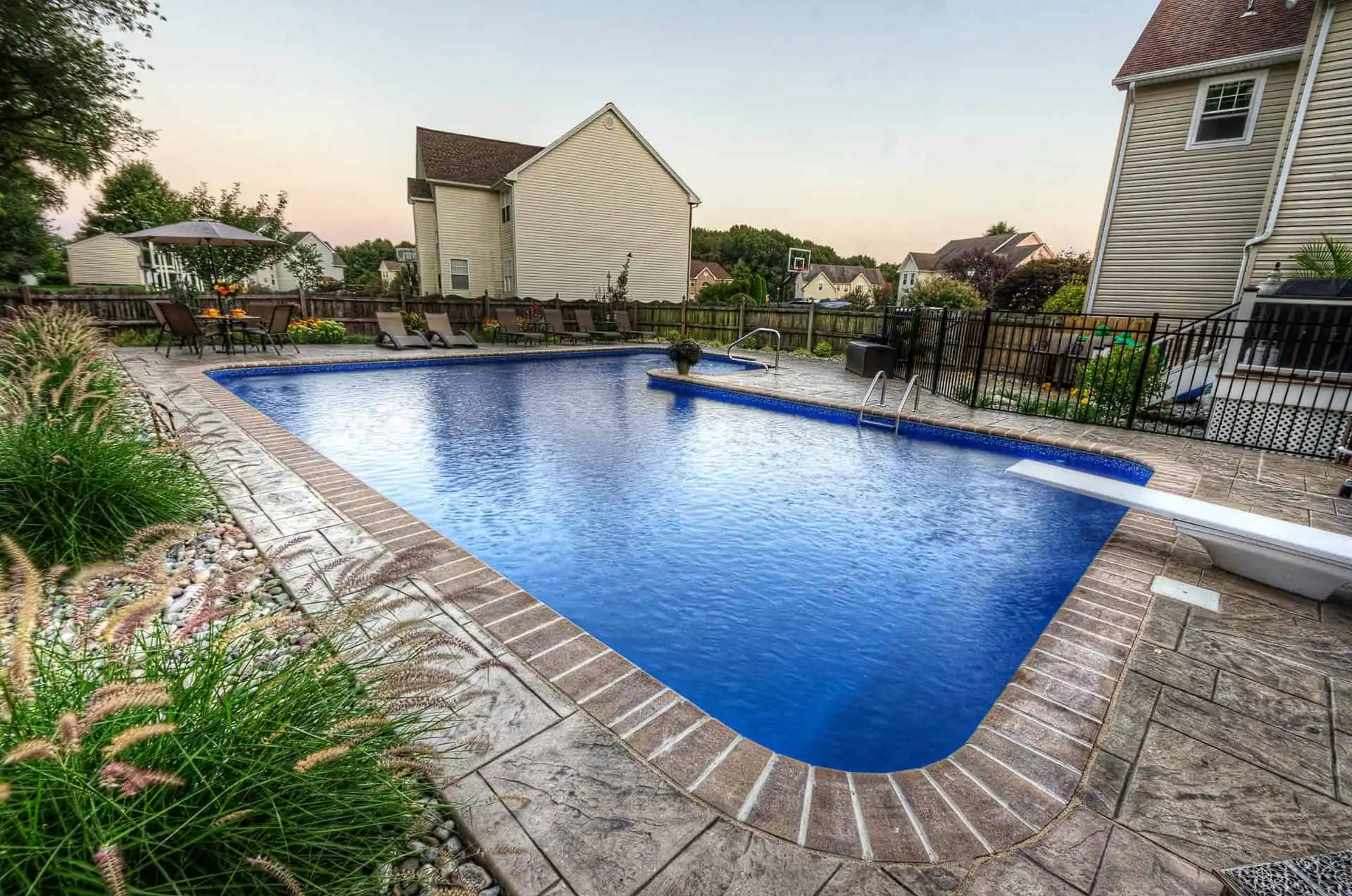 A large, rectangular backyard swimming pool with a diving board, surrounded by stamped concrete, lounge chairs, and landscaped plants, located next to suburban houses in a residential neighborhood at dusk.