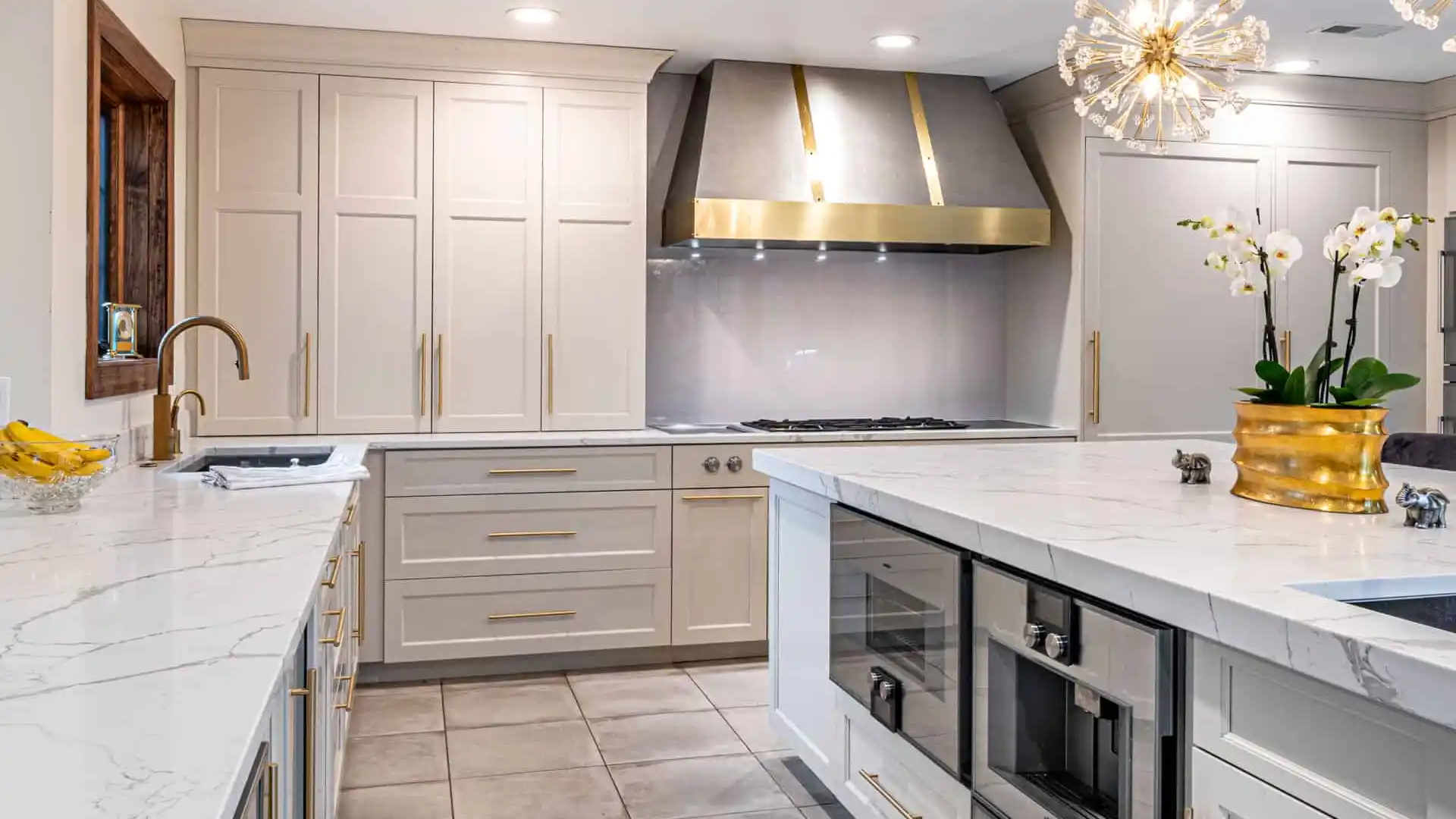 A modern kitchen featuring white cabinets, a marble countertop, a large stainless steel range hood, and a central island with built-in appliances. A chandelier hangs from the ceiling, and a potted orchid decorates the island—a perfect indoor complement to your outdoor living space in Delaware County PA.