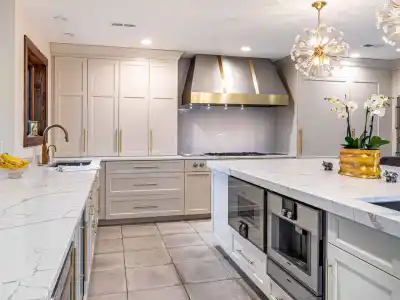 A modern kitchen featuring white cabinets, a marble countertop, a large stainless steel range hood, and a central island with built-in appliances. A chandelier hangs from the ceiling, and a potted orchid decorates the island—a perfect indoor complement to your outdoor living space in Delaware County PA.