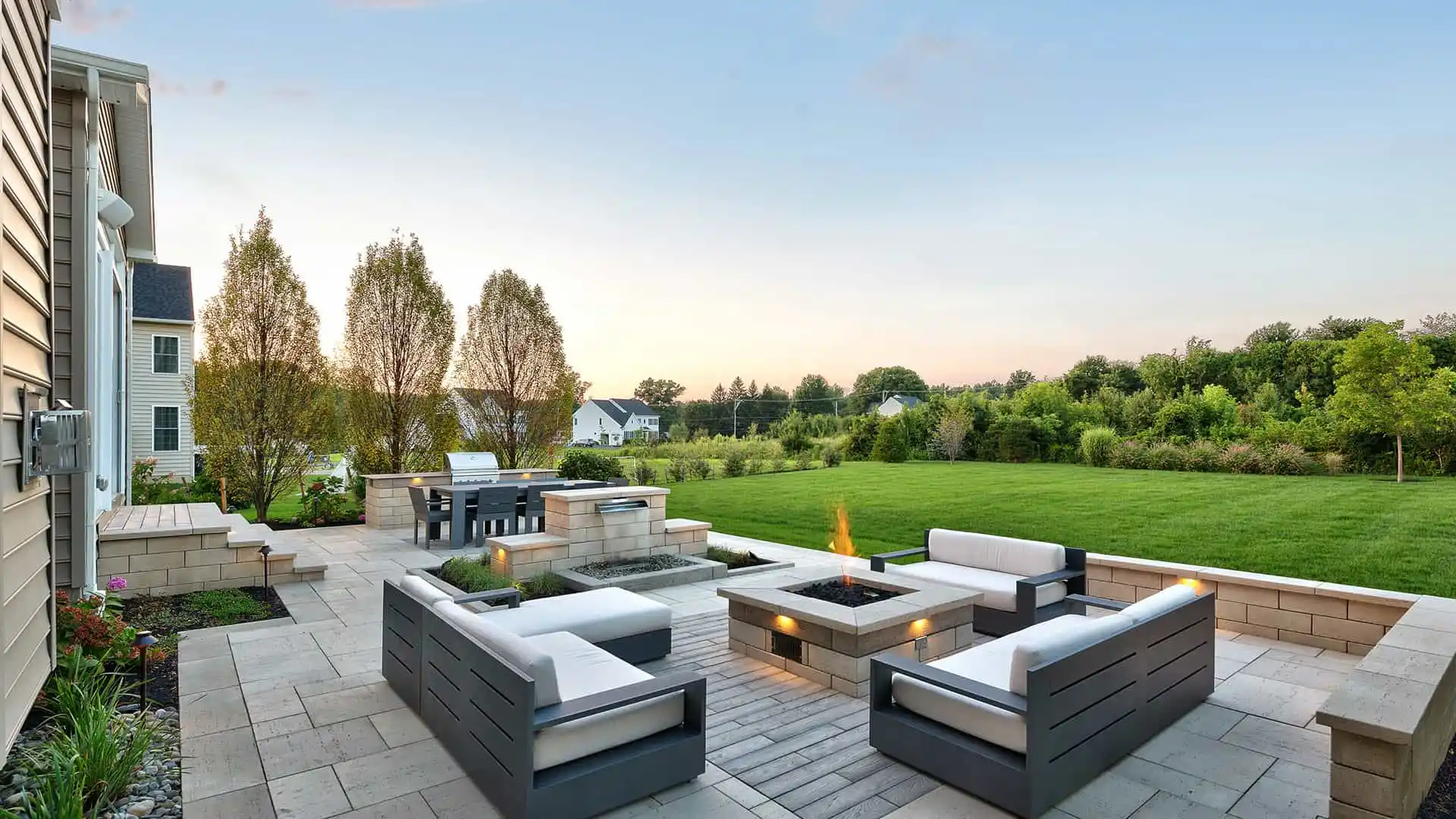 A spacious patio with modern outdoor seating, centered around a square fire pit, exemplifies premier outdoor living in Delaware County, PA. The area features a barbecue and overlooks a large, green lawn bordered by trees. The sky is clear with a hint of sunset.