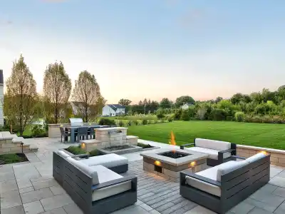 A spacious patio with modern outdoor seating, centered around a square fire pit, exemplifies premier outdoor living in Delaware County, PA. The area features a barbecue and overlooks a large, green lawn bordered by trees. The sky is clear with a hint of sunset.
