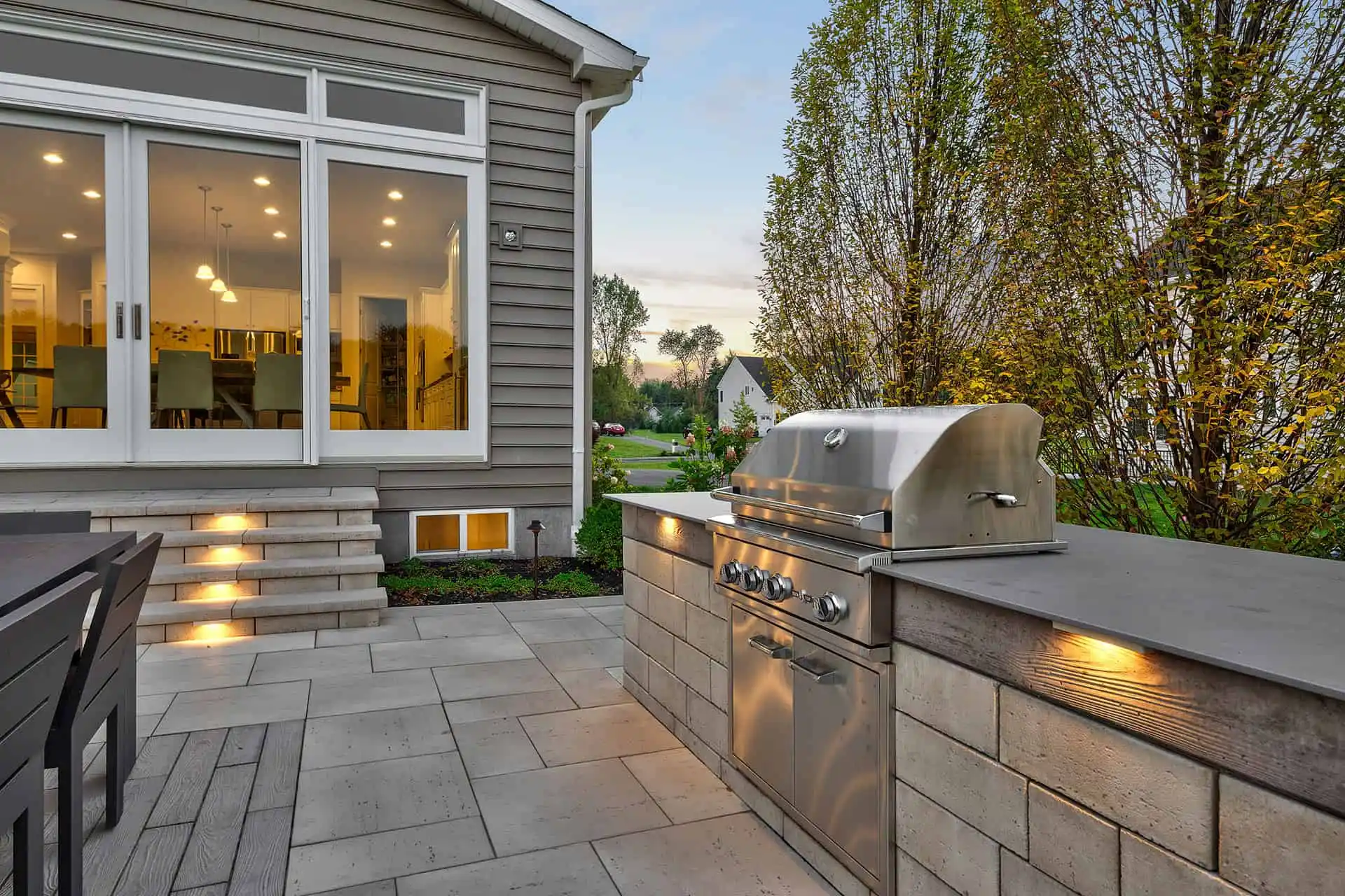 Modern outdoor patio with built-in stainless steel grill, stone countertops, and dining area. Steps lead to a house with large windows and warm interior lights, surrounded by trees and landscaping.