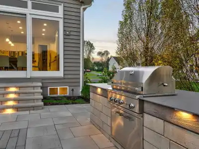 Modern backyard patio in Delaware County, PA, featuring a stainless steel grill on a stone countertop. Warm lights accentuate the grill area. Nearby, a dining table with chairs awaits. Large glass doors lead to an illuminated interior space with a dining room, perfect for outdoor living enthusiasts.