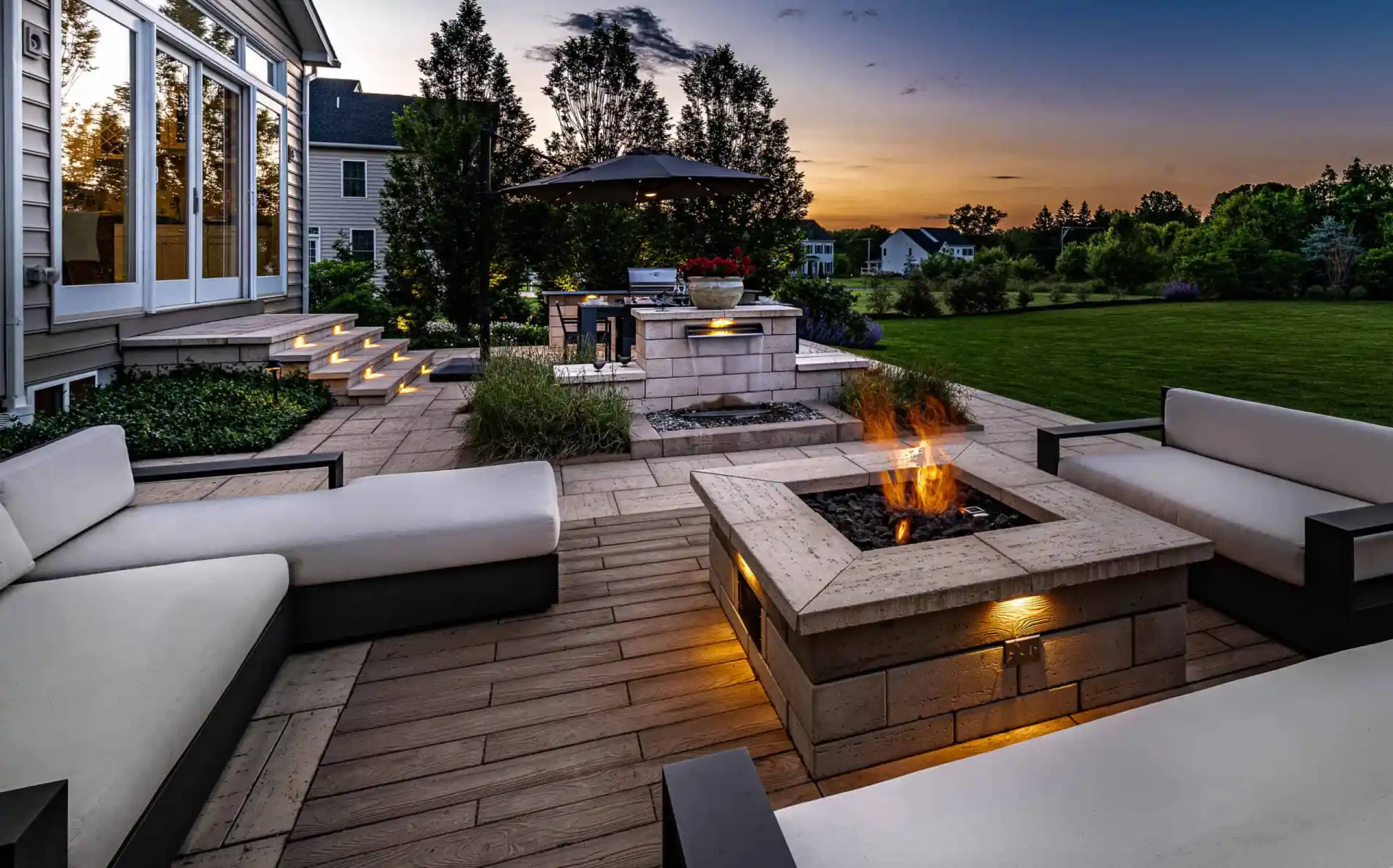 A modern outdoor patio at sunset, showcasing exquisite outdoor living in Delaware County, PA, features white cushioned seating around a lit square fire pit. Illuminated steps lead to a house, with an umbrella shading the grill area. Trees and a lawn frame the scene under a colorful sky.