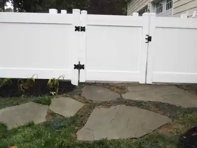 White vinyl fence with black hinges beside a beige house, featuring two gates on a blue-gray stone pathway lined with mulch and plants. Enhance your outdoor living in Delaware County, PA, with professional landscaping services. Trees add a natural backdrop to the serene scene.