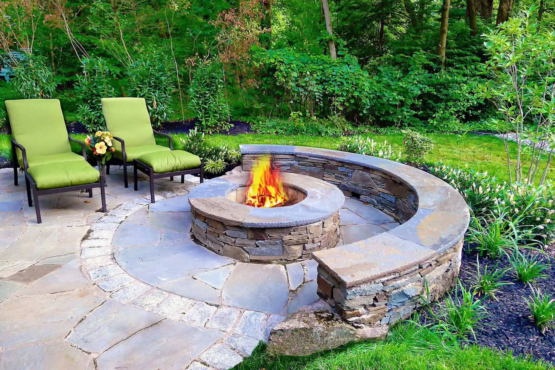 A round stone fire pit with a curved stone bench and two green cushioned lounge chairs sits on a stone patio, surrounded by lush greenery and trees in a backyard setting.