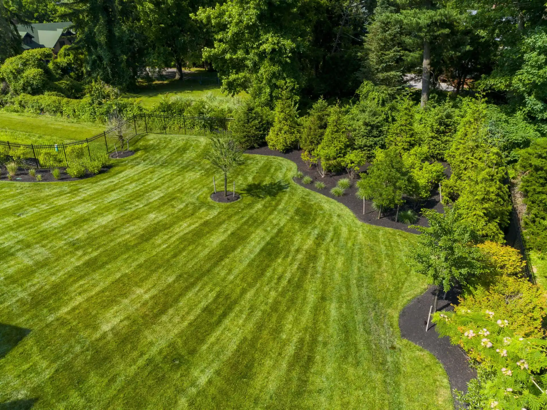 A well-maintained lawn with visible mowing stripes, bordered by neatly mulched flower beds and young trees, surrounded by dense green shrubs and tall trees in the background.