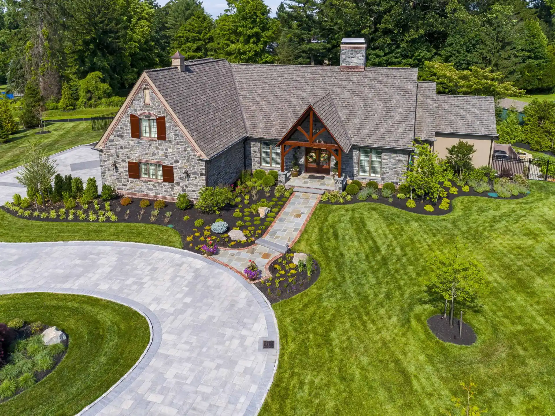 Aerial view of a large stone house with a gabled roof, landscaped gardens, a curved driveway, and well-manicured lawn surrounded by lush green trees and shrubs.