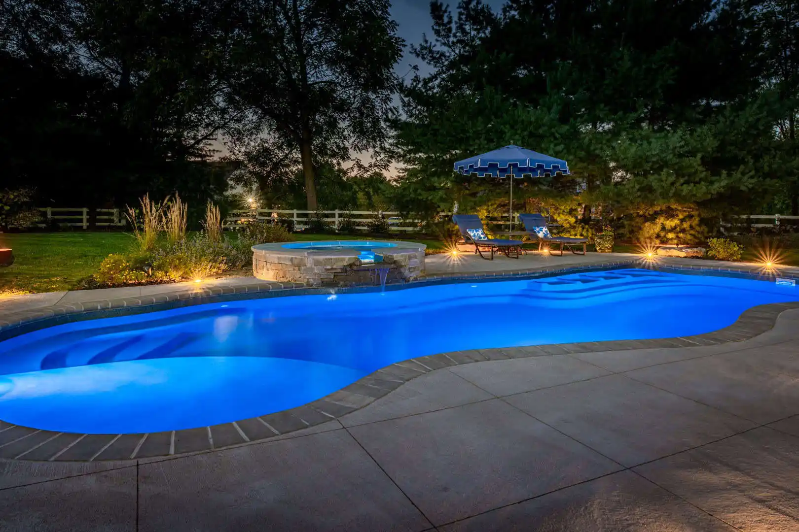 A lit-up backyard pool at dusk, with glowing blue water and a connected circular hot tub, epitomizes outdoor living in Delaware County PA. Two lounge chairs and a striped umbrella on the right are beautifully framed by trees and soft landscape lighting, showcasing expert landscaping services in Delaware County PA.