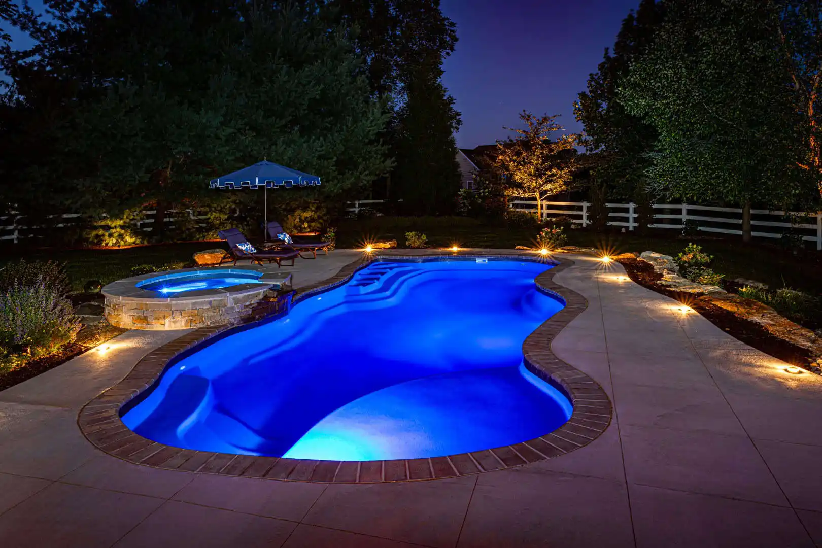 A backyard scene at night features a glowing blue swimming pool with an attached circular hot tub. Lounge chairs and a large umbrella adorn the deck, crafted by expert hardscaping services in Delaware County, PA. Trees and a white fence surround the area, lit by subtle landscape lighting.