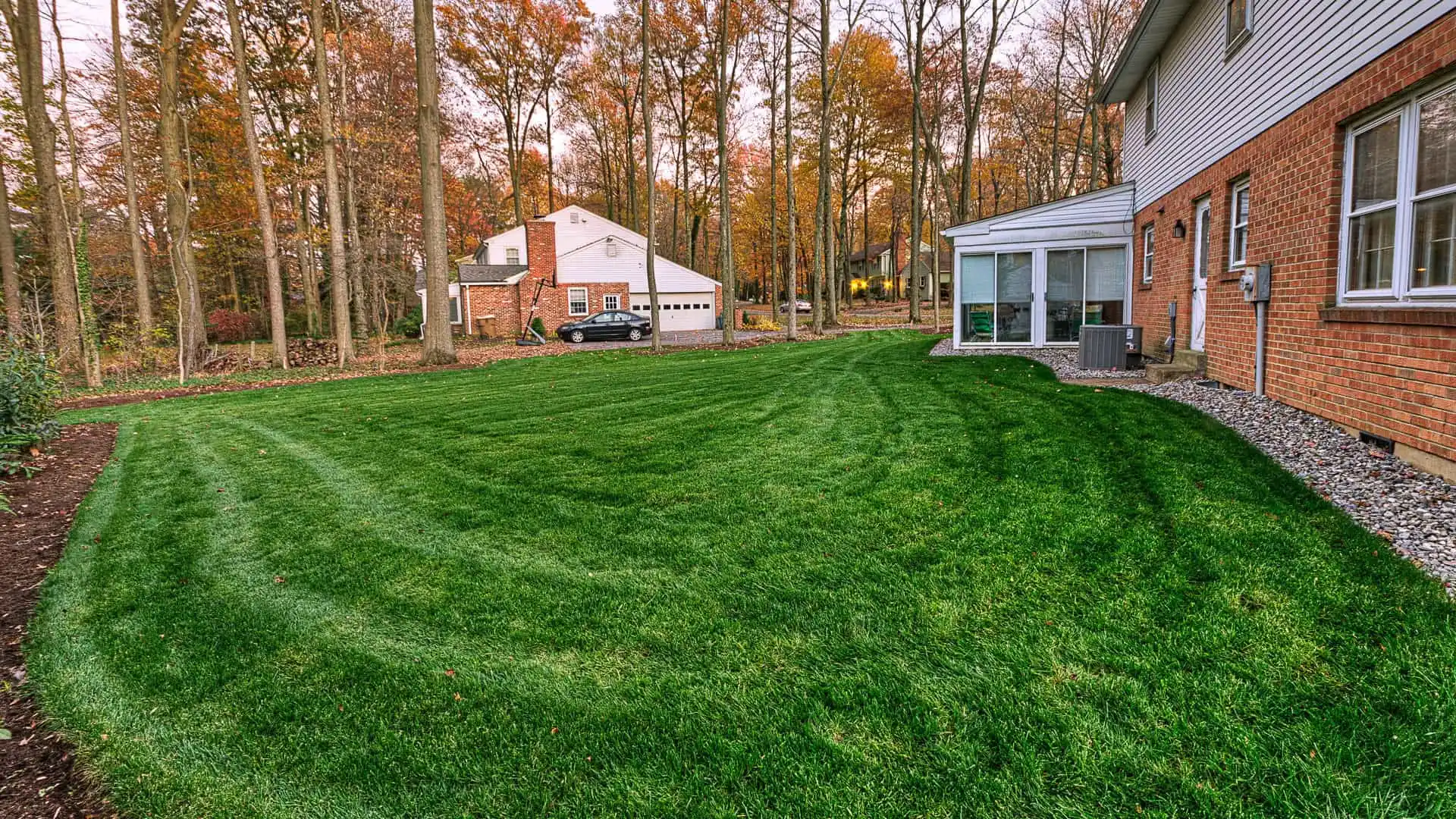 A neatly mowed grassy backyard enhances the charm of a red brick house on the right, with a glass-walled sunroom extending from it. Surrounded by autumn trees, another house with a garage offers ample outdoor living space in Delaware County, PA. A car is parked in front of the garage.
