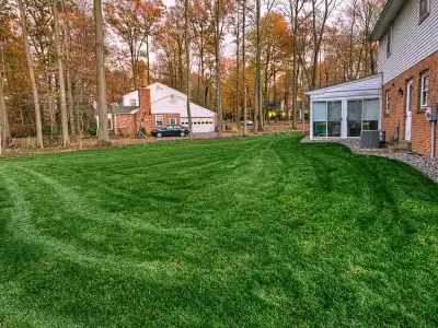 A neatly mowed grassy backyard enhances the charm of a red brick house on the right, with a glass-walled sunroom extending from it. Surrounded by autumn trees, another house with a garage offers ample outdoor living space in Delaware County, PA. A car is parked in front of the garage.