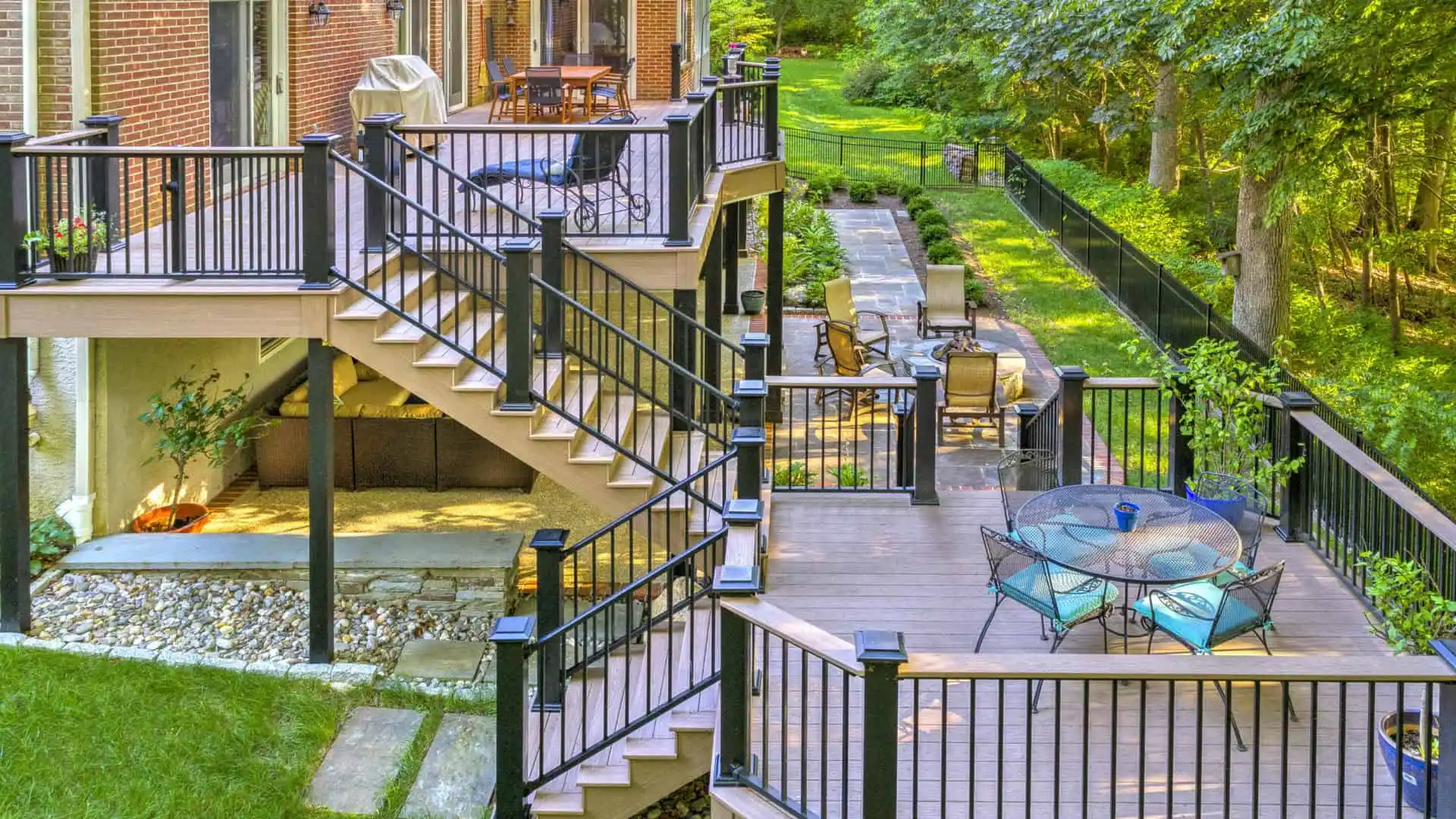 A multi-level deck attached to a brick house features outdoor furniture, including tables and chairs. The deck, crafted with expert hardscaping services in Delaware County, PA, overlooks a green backyard with a pathway lined by a fence and trees. Steps connect the different levels of the deck.