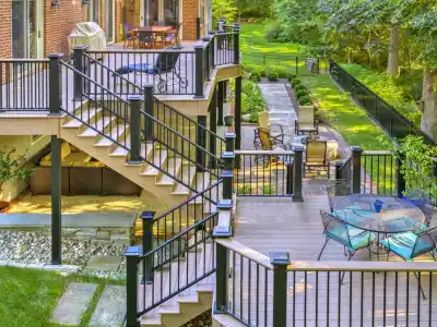 A multi-level deck attached to a brick house features outdoor furniture, including tables and chairs. The deck, crafted with expert hardscaping services in Delaware County, PA, overlooks a green backyard with a pathway lined by a fence and trees. Steps connect the different levels of the deck.