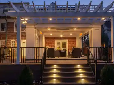 A well-lit patio attached to a brick house features a pergola with string lights, perfect for outdoor living in Delaware County, PA. Steps with embedded lights lead up to the area where cushioned chairs and a table are arranged under the pergola.
