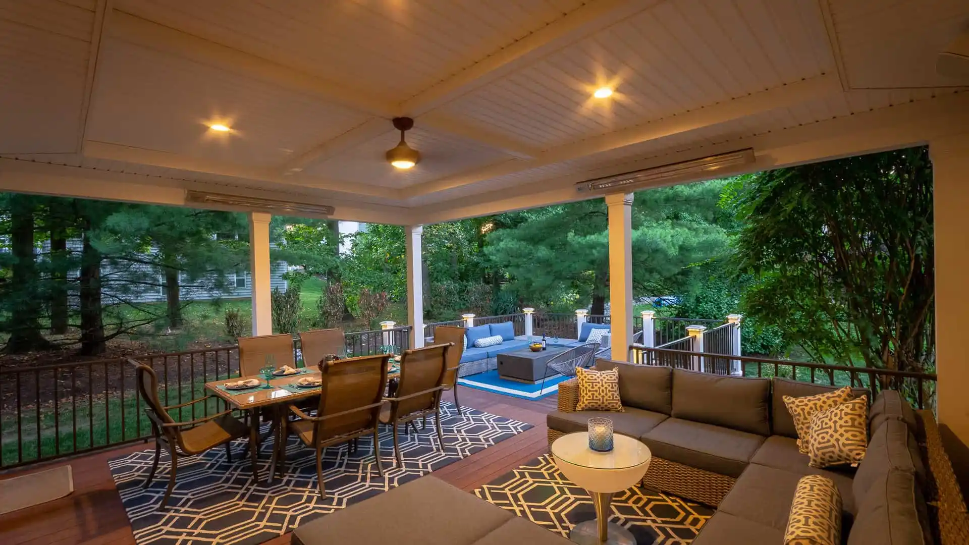 A cozy outdoor patio in Delaware County, PA, enhanced by expert outdoor living design. It features a wooden ceiling with recessed lighting, a corner sectional sofa, a dining table with chairs, and a view of trees. A patterned rug and cushions add warmth, with a small fire pit table visible in the background.