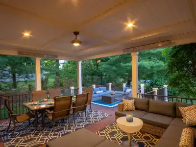 A cozy outdoor patio in Delaware County, PA, enhanced by expert outdoor living design. It features a wooden ceiling with recessed lighting, a corner sectional sofa, a dining table with chairs, and a view of trees. A patterned rug and cushions add warmth, with a small fire pit table visible in the background.