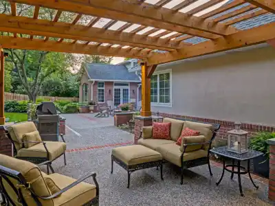 A cozy outdoor patio in Delaware County, PA, features a wooden pergola with comfortable cushioned seating around a small table on a paved area. A barbecue grill is nearby, surrounded by well-maintained landscaping services and trees in the background.