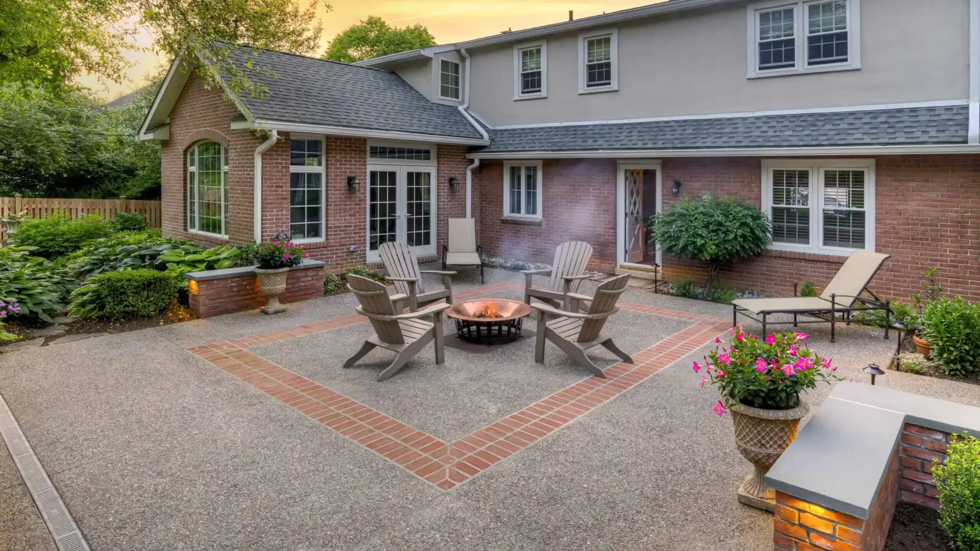 A spacious backyard patio in Delaware County, PA, showcases a central fire pit surrounded by wooden chairs. Bordered with a brick design and complemented by lounge chairs and potted plants, this outdoor living space is set against a two-story brick and siding house under a sunset sky.