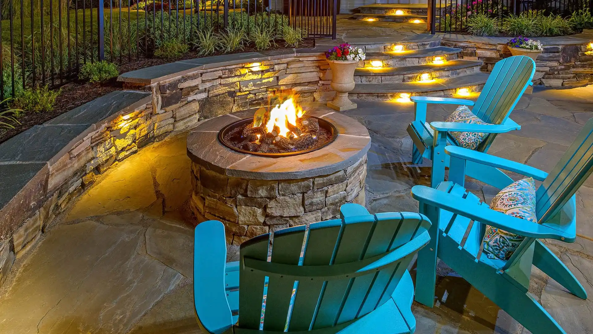 A cozy outdoor patio, crafted with expert hardscaping services in Delaware County, PA, features a stone fire pit with a bright flame. Surrounding the pit are turquoise Adirondack chairs. In the background, stone steps with glowing lights lead upward, flanked by plants and a stone wall.