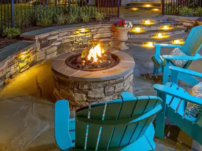 A cozy outdoor patio, crafted with expert hardscaping services in Delaware County, PA, features a stone fire pit with a bright flame. Surrounding the pit are turquoise Adirondack chairs. In the background, stone steps with glowing lights lead upward, flanked by plants and a stone wall.